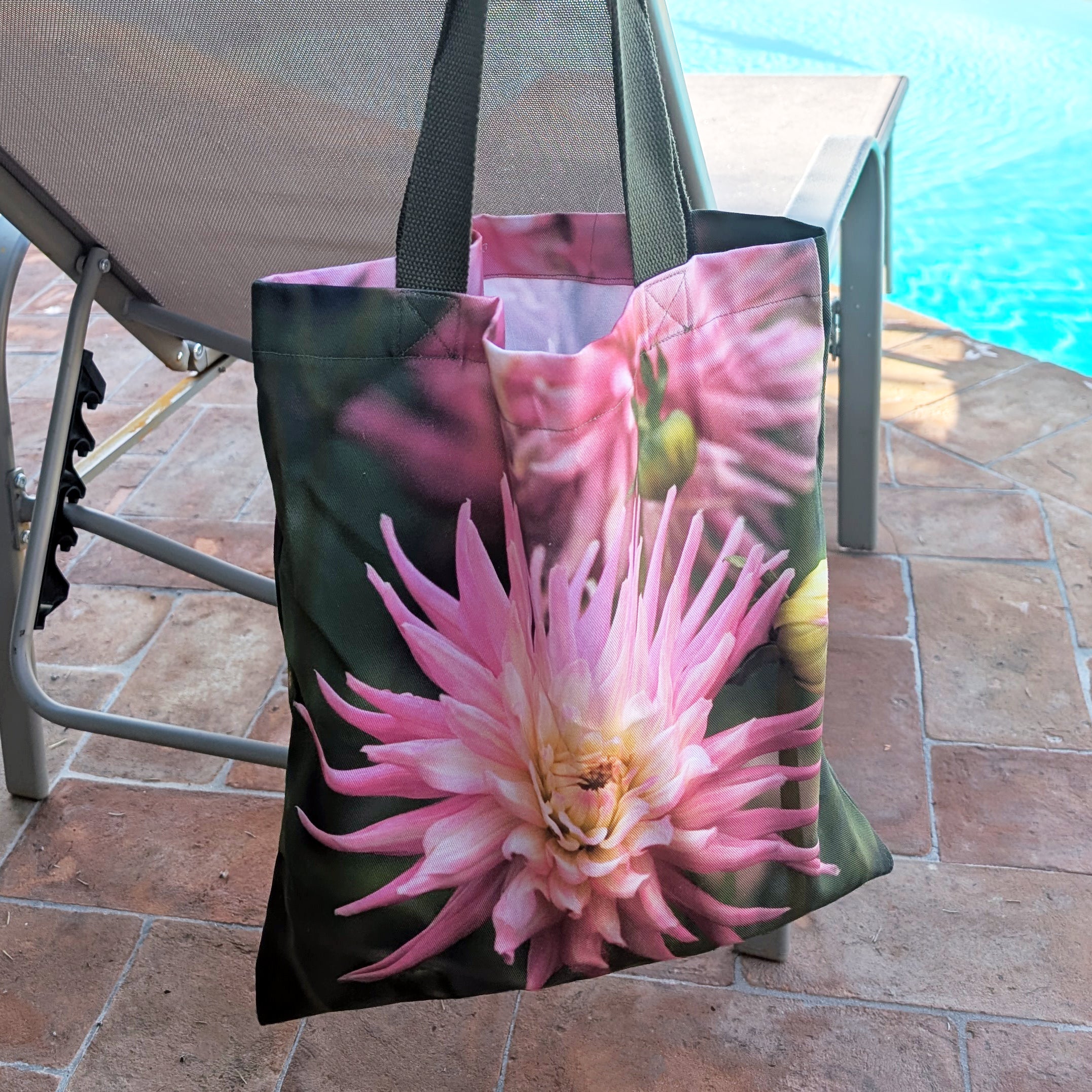 Tote bag with pink dahlia design on a patio chair near a pool.