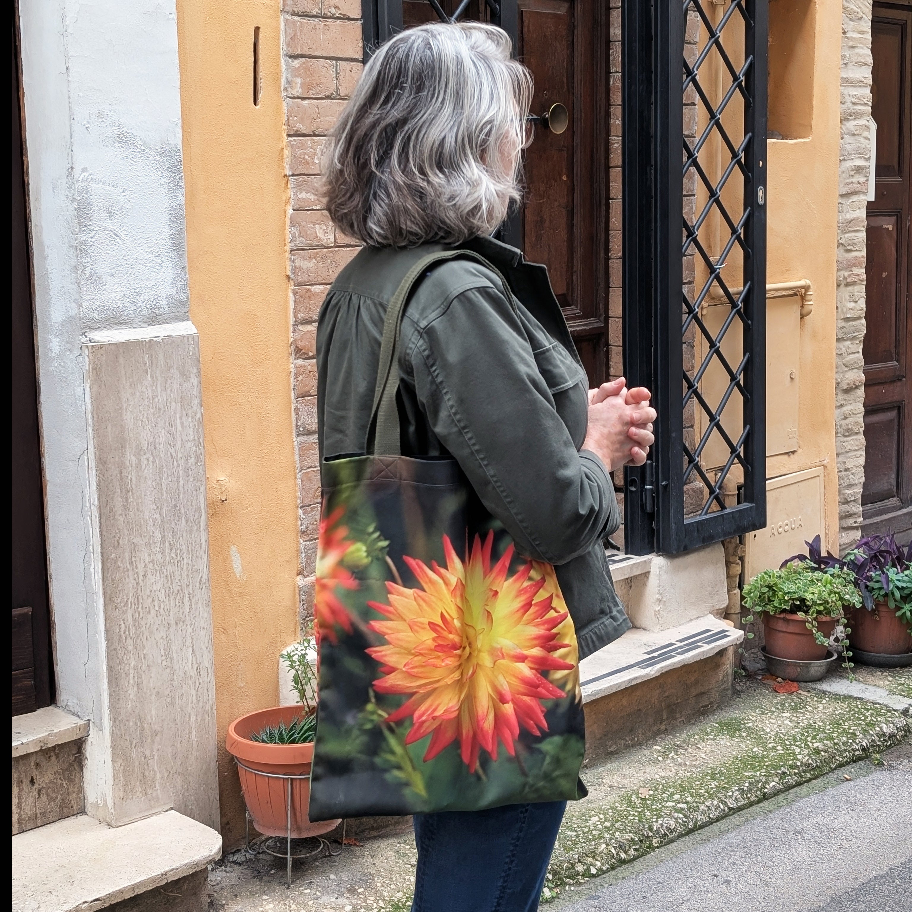 Person holding a Dahlia Days tote bag with a colorful orange dahlia design, standing outside a building in Italy.